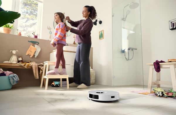 Robot aspirapolvere Bosch Spotless in funzione sul pavimento di un bagno, mentre una donna pettina i capelli a una bambina