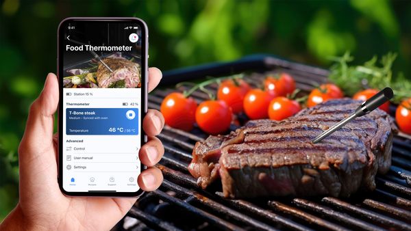 Roka s pametnim telefonom, ki prikazuje, kako aplikacija Bosch Home Connect spremlja postopek kuhanja zrezka t-bone. V ozadju je na žaru videti govedino s sondo za meso.