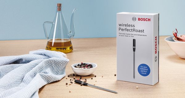 Slika zaprte embalaže brezžične sonde za meso Bosch PerfectRoast, ob njej je na lesenem pultu še odprta embalaža, obdana s sestavinami in kuhinjsko brisačo.
