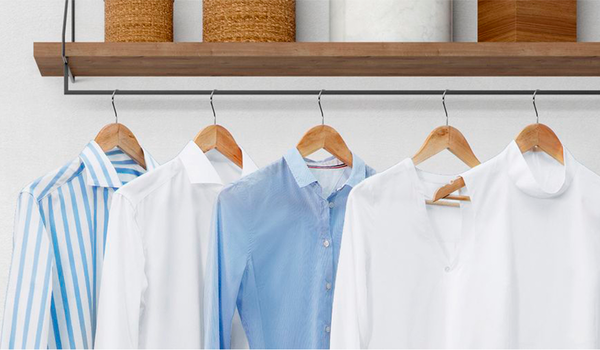 Mehrere frisch geglättete Hemden und Blusen hängen nebeneinander auf Holzkleiderbügeln unter einem Regal mit Körben – die Kleidung ist dank der Bosch Waschmaschine mit Dampffunktion glatt und gepflegt.