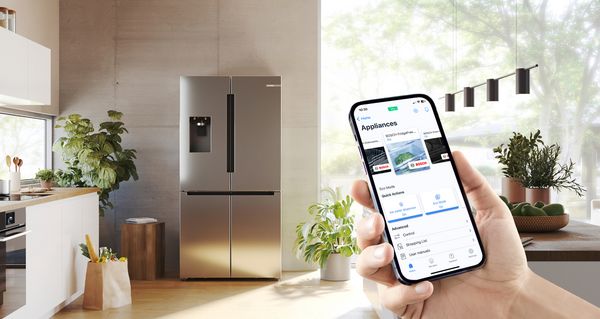 Een hand houdt een smartphone vast die de inhoud van een koelkast laat zien, die zichtbaar is in de lichte keuken op de achtergrond.
