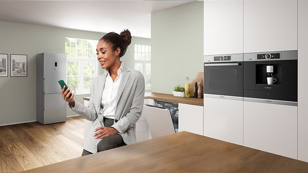 Een vrouw in een moderne keuken zit aan een houten tafel, houdt een smartphone vast, met een koelkast en apparaten 