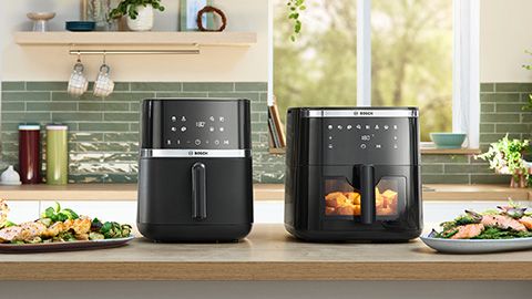 Eine Mutter und ihre Tochter beobachten durch das Fenster einer Bosch Heißluftfritteuse Serie 6 erwartungsvoll, wie Lebensmittel in dem Gerät garen. Die Küche ist hell und modern.