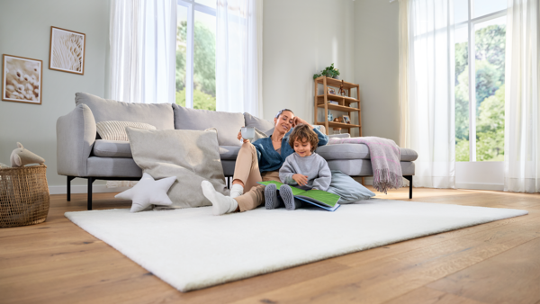 Eine Frau sitzt mit einem Kind in einem Wohnzimmer auf einem weißen Teppich und sie schauen gemeinsam ein Buch an.