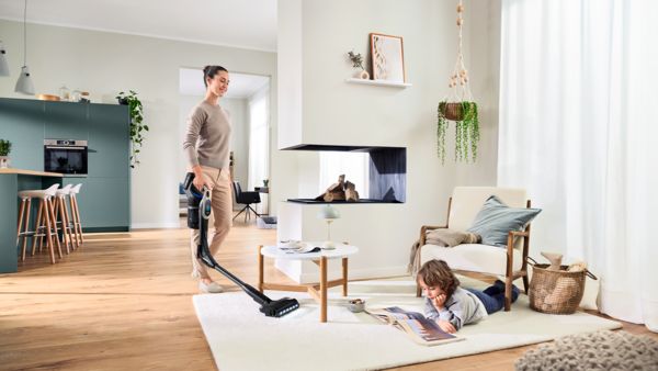 Ein Kind liegt bäuchlings auf einem weißen Wohnzimmerteppich und betrachtet einen Bildband während eine Frau mit dem Bosch Unlimited 10 mit knickbarem Saugrohr den Teppich saugt.