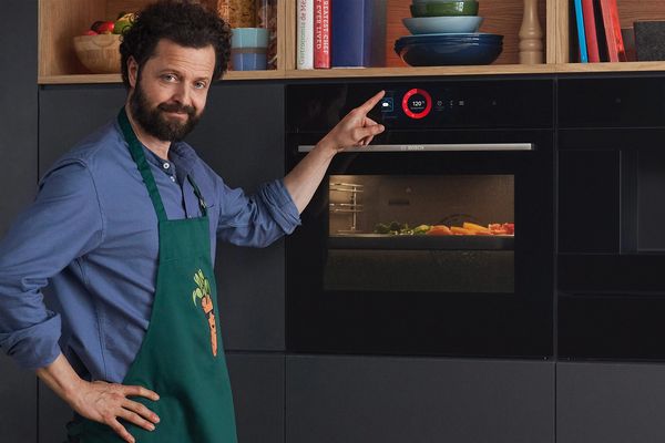 Mann zeigt auf das Bedienfeld eines modernen Ofens mit Gemüse darin in einer Küchenszene.