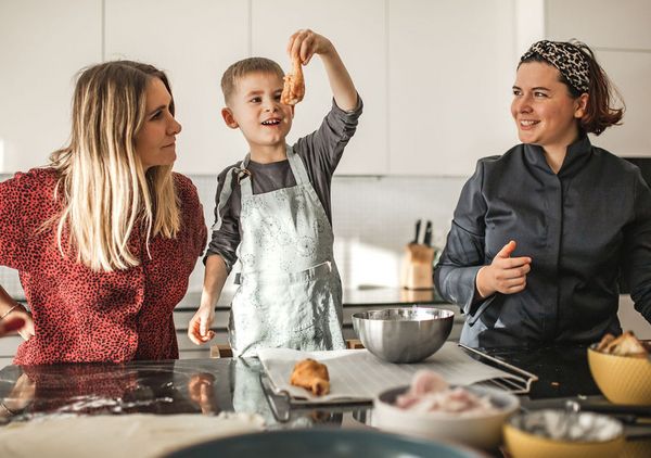 Kind hält ein Lebensmittel in der Küche, zwei lächelnde Frauen bereit zum gemeinsamen Kochen.