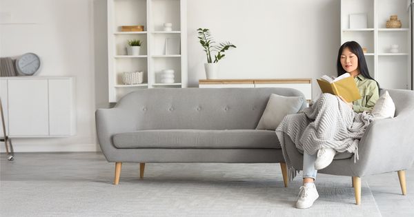 A person sits on a gray couch, wrapped in a blanket, reading a book in a minimalist living room.