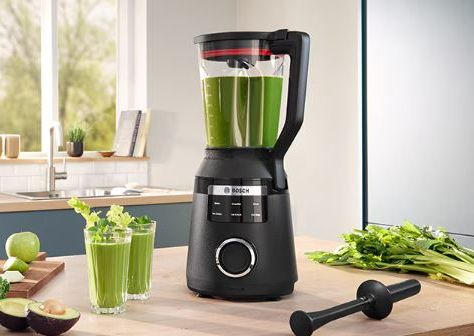 Una batidora Bosch negra sobre la encimera de la cocina, llena de batido verde, junto a dos vasos de bebida verde y tallos de apio.