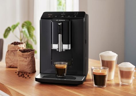 Una elegante cafetera negra prepara café espresso, con dos tazas de café y bolsas de granos de café sobre una encimera de madera.