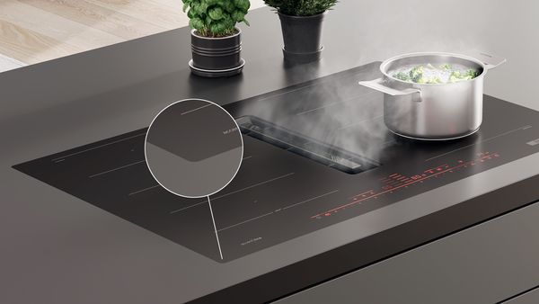 Matzwarte kookplaat met afzuiging in zwart werkblad. Een cirkel licht uit hoe de kookplaat in het werkblad verwerkt is: vlak geïntegreerd waardoor de kookplaat bijna één is met het werkblad.   