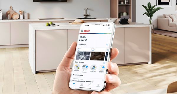 Ένα χέρι το οποίο κρατάει ένα smartphone σε πρώτο πλάνο, με μια όμορφη εικόνα κουζίνας στο φόντο.