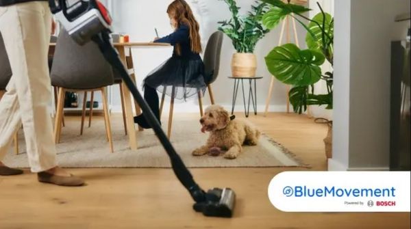 Een persoon stofzuigt in een knusse kamer, terwijl een kind aan tafel zit te schetsen en een hond in de buurt toekijkt. Groene planten versterken de ruimte.