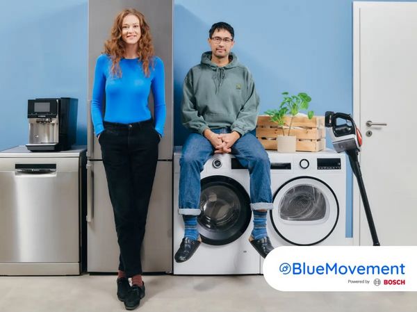 Een vrouw in een blauw topje staat naast een man in een hoodie, vlakbij moderne apparaten, waaronder een koelkast, wasmachine en een plant.