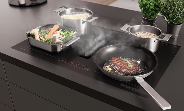 Strak keukenblad met inductiekookplaat, sissende steak, stoomende groenten, pasta en champignons in pannen.