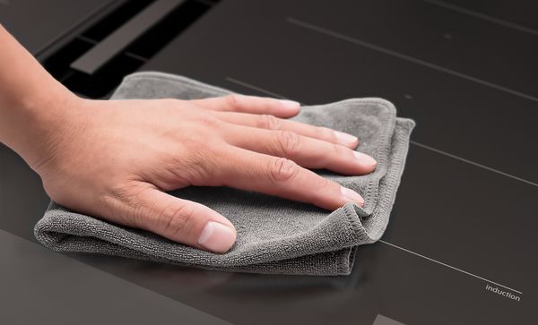 Hand met grijze doek maakt strak zwart oppervlak van inductiekookplaat schoon.