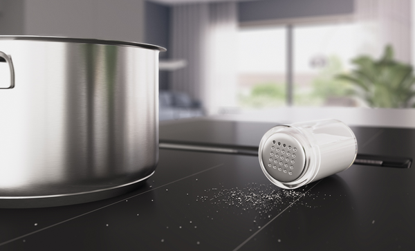 Gros plan d'une sali&egrave;re renvers&eacute;e sur la table de cuisson aspirante Series 8 au design mat noir carbone. Du sel sur la surface, &agrave; c&ocirc;t&eacute; d'une casserole chrom&eacute;e.