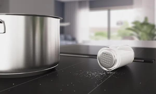 Крупный план упавшей солонки на варочной панели Series 8 с интегрированной вытяжкой в матовом угольно‑чёрном исполнении. Соль рассыпана по поверхности рядом с хромированной кастрюлей.