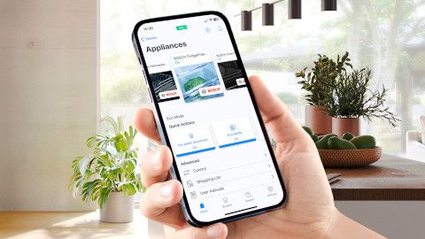 Pralni stroj Bosch v sodobni kopalnici z roko v ospredju, ki drži pametni telefon, na katerem je odprta aplikacija Home Connect