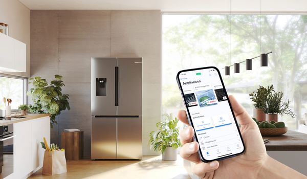 Une main tient un smartphone affichant une application de r&eacute;frig&eacute;rateur intelligent avec un fond de cuisine moderne.