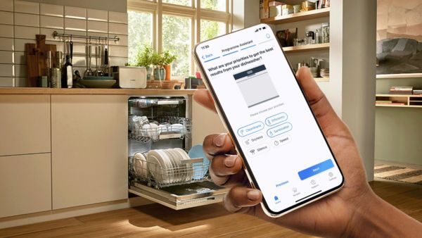 A hand holds a smartphone displaying a dishwasher settings questionnaire, next to an open dishwasher filled with clean dishes in a bright kitchen.