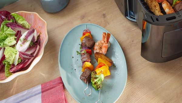 Spiedini di carne, verdure e gamberi serviti su un piatto azzurro accanto a una friggitrice ad aria Bosch e a un&rsquo;insalata