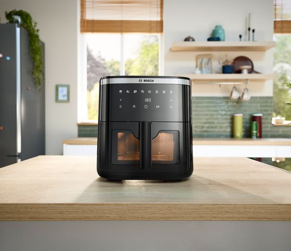 Eine elegante schwarze Heißluftfritteuse von Bosch steht auf einer Holzarbeitsplatte in einer hellen Küche und verfügt über digitale Bedienelemente und ein transparentes Fenster.