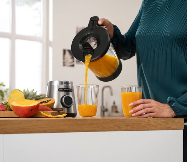 Eine Frau gießt frischen Orangen-Smoothie in zwei Gläser, umgeben von Früchten und einem Mixer auf einer Küchenarbeitsplatte.