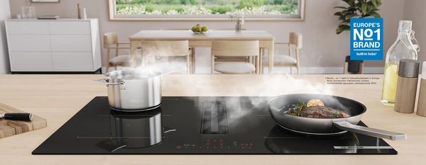 Bosch-kookplaat met geïntegreerde ventilatiemodule. Een braadpan met een biefstuk en een kookpan op de kookplaat, eetruimte met eettafel op de achtergrond.