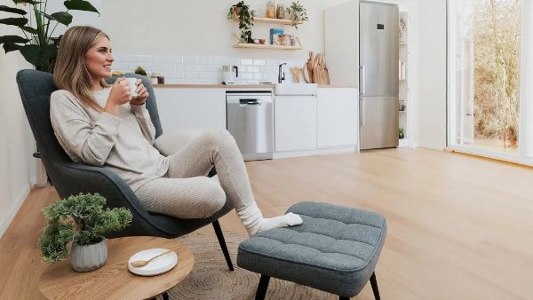 Um espaço de estar aconchegante com uma pessoa vestindo roupas confortáveis a beber chá, rodeada por uma mesa de café de madeira e uma cozinha moderna.   