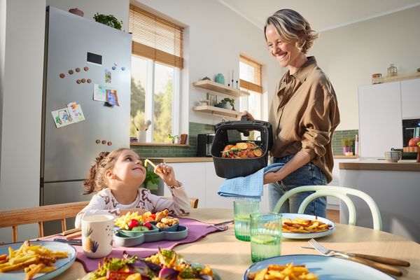 Kobieta i dziecko w kuchni air fryer na blacie w kuchni, obok potrawy na talerzach