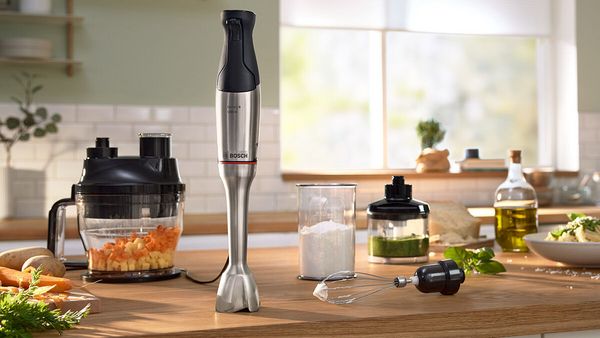 Ręczny blender marki Bosch stoi na kuchennym blacie, otoczony pokrojonymi warzywami, robotem kuchennym, przyprawami i oliwą z oliwek. Jest to eksponat przedstawiający sprzęt do gotowania. 