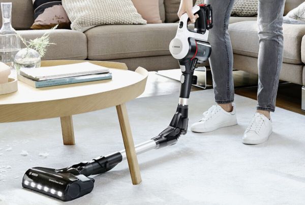 A woman uses a cordless vacuum to clean up a mess on a cream rug in light, bright living room.