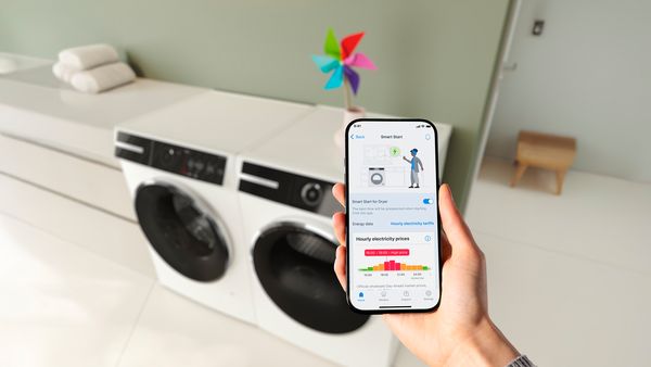 Een wasmachine en droogkast staan op de achtergrond, met op de voorgrond een smartphone die het uurtarief van de elektriciteit toont in de Bosch app.