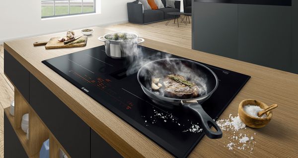 Inductiekookplaat met geïntegreerde afzuigkap in houten keukeneiland tijdens het koken van vis en groenten met zichtbare stoom