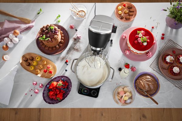 Sul piano di lavoro della cucina sono esposti uno sbattitore elettrico, vari dessert, frutti di bosco freschi e utensili da forno, creando una vivace scena di pasticceria. 