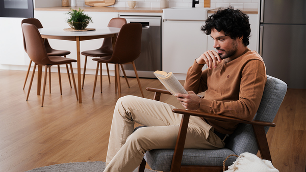 Un homme assis dans un fauteuil en train de lire un livre, dans un espace cuisine‑salle &agrave; manger moderne avec un parquet en bois et des chaises brunes.