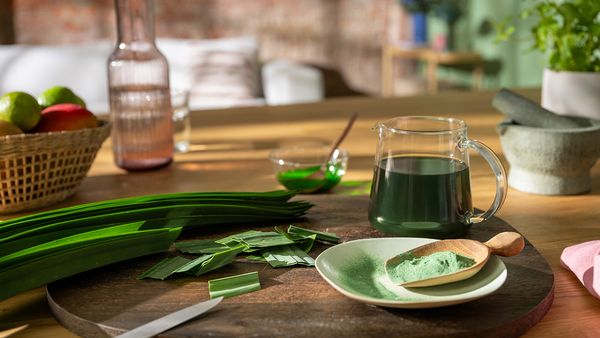 Auf einem Tablett liegen Pandan-Blätter neben einem Teller mit Pandan-Pulver und einer kleinen Kanne gefüllt mit Pandan-Saft.