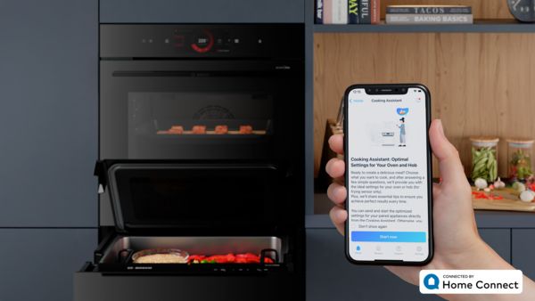 Eine Person hält ein Smartphone mit der geöffneten Bosch Home Connect Kochassistent-App in der Hand, während im Hintergrund der accent line Dampfgarer geöffnet ist.