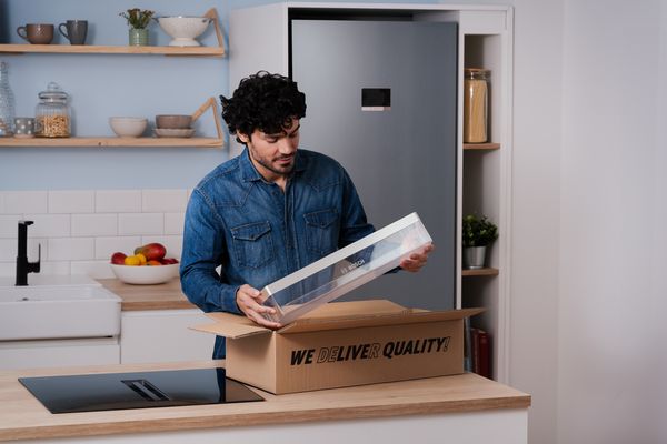 En man i ett kök packar upp en leverans. Han håller i en ny kylskåpslåda från Bosch som han lyft ur en kartong med texten "WE DELIVER QUALITY!" på köksbänken.