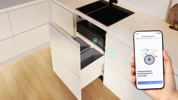 Ein offener Schubladen in einer hellen Einbauküche mit einem Smartphone in der Hand, das den Saub- und Wischroboter aus der Home Connect App heraus steuert.