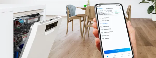 יד מחזיקה בטלפון חכם שמציג תוכניות לשימוש במדיח כלים. המדיח פתוח ומכיל כלים, ברקע כסאות מעץ וצמחייה.   