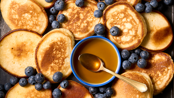 Viele kleine Pfannenkuchen mit Blaubeeren und einer kleinen weißen Schale mit Ahornsirup, in der ein silberner Löffel liegt.