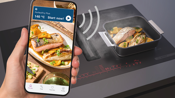 Ein Smartphone wird von einer Hand gehalten und zeigt in der BetterFood App ein Gericht mit Details wie Temperatur und Sensoreneinstellung, vor einem verschwommenen Küchen-Hintergrund.