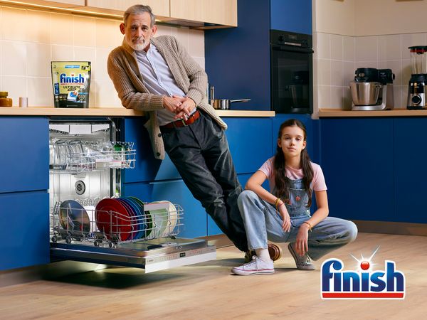Adult and child in kitchen next to open Dishwasher with Finish pack on top