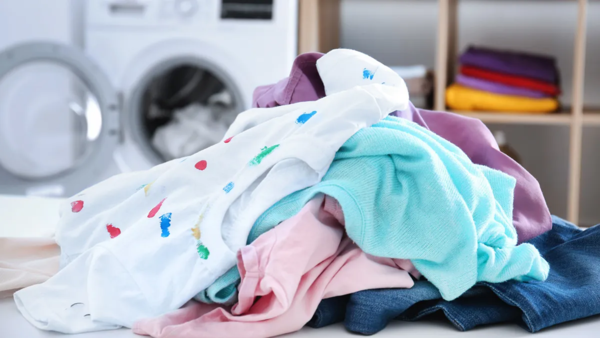 A pile of colorful laundry sits in front of an open washing machine, with shelves of neatly folded laundry in the blurred background.