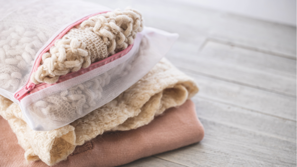 A close-up of a stack of folded laundry, possibly towels or blankets, with a mesh laundry bag containing a fluffy, light-colored item on top, all resting on a light-colored wooden surface.