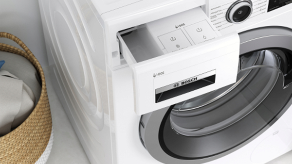 A close-up view of the detergent dispenser drawer of a white front-loading washing machine, partially pulled out, with a laundry basket visible on the left.