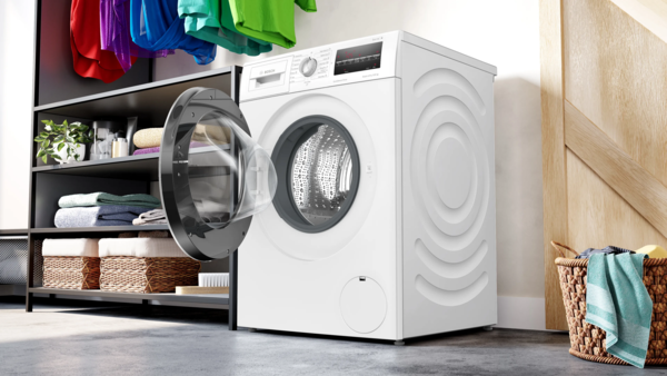 A white Bosch front-load washing machine is centered in a laundry room, with shelving holding folded clothes and storage boxes to the left.