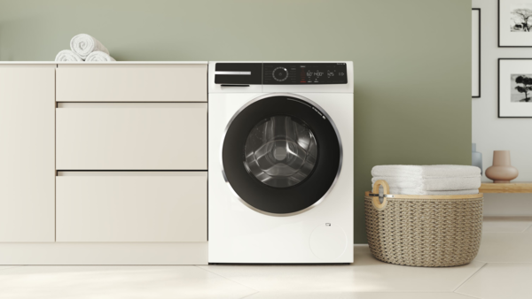 Eine Waschmaschine an der Seite einer Küche, links daneben ein weißer Stuhl. In der rechten Bildecke sehen wir einen Backofen und eine Arbeitsplatte mit einer Pflanze.